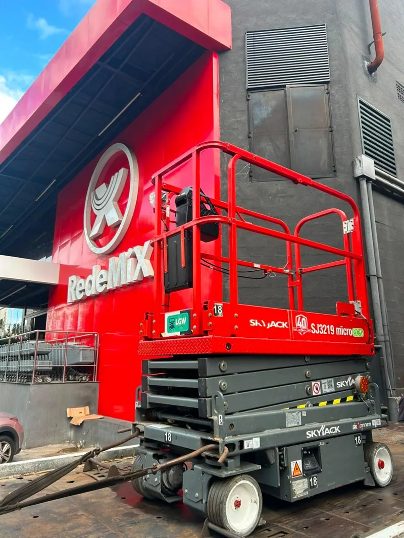 Plataforma tesoura cinza com guarda-corpo vermelho em frente à fachada vermelha de um supermercado