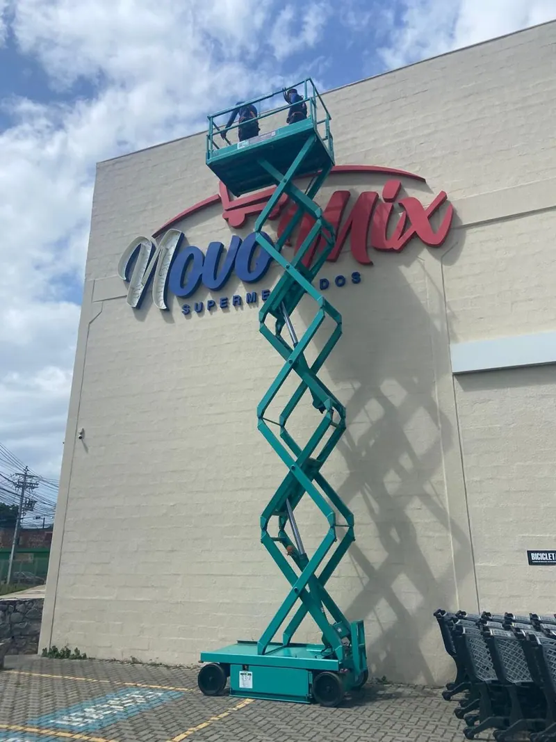 Plataforma tesoura verde-água estendida até o alto, em frente à fachada de um supermercado