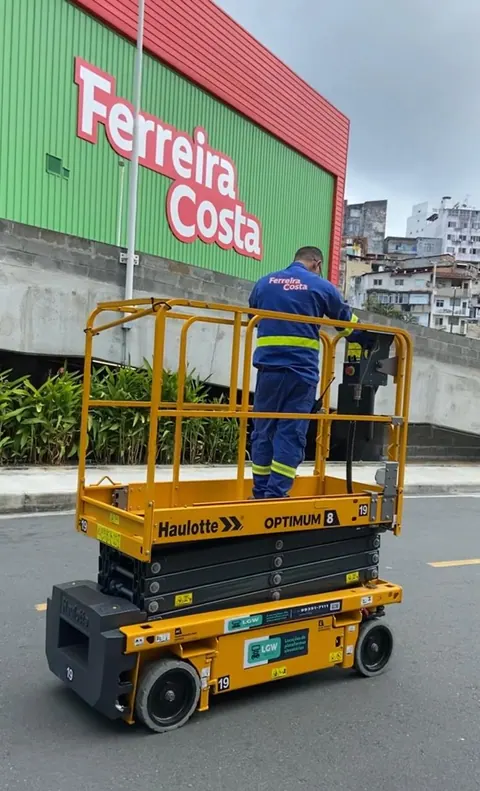 Plataforma tesoura amarela com um operador de uniforme azul, em frente a uma loja verde e vermelha